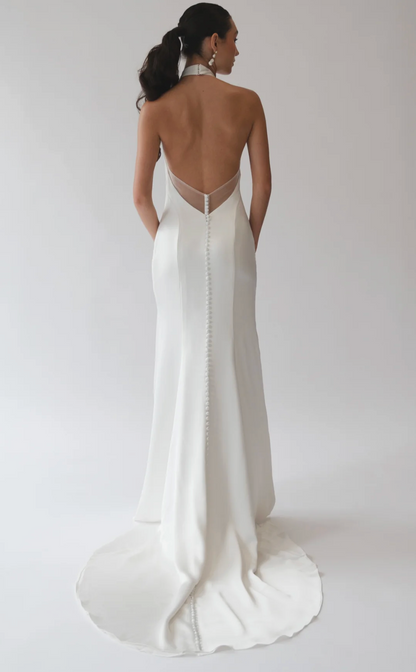A woman shown from the back wearing a fitted white bridal gown with an open back, sheer mesh panel at the neckline, and a long train on a light gray studio background.
