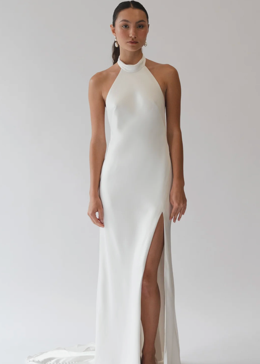 A model wearing a floor-length white halter gown with a high thigh slit, photographed against a seamless light gray studio background.