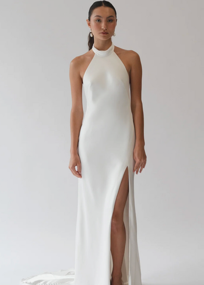 A model wearing a floor-length white halter gown with a high thigh slit, photographed against a seamless light gray studio background.