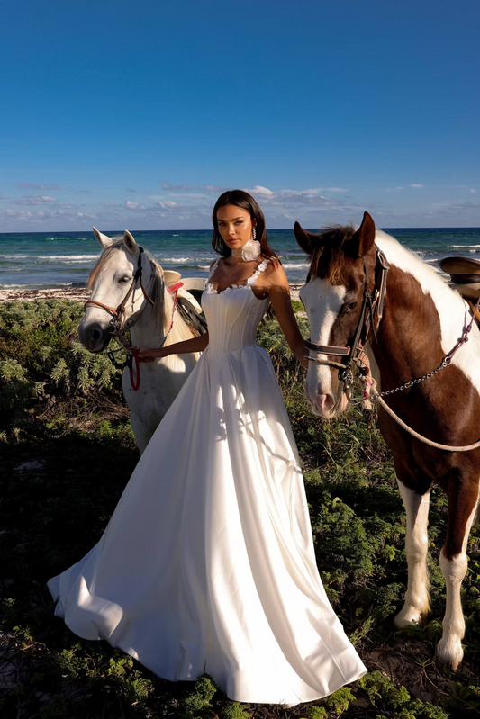 A woman in a white wedding gown stands outdoors between two horses on a beach, with ocean, sky, and low shrubs in the background.