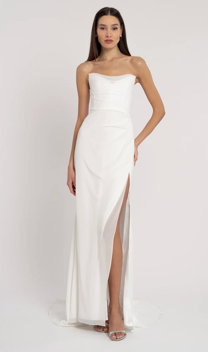A woman modeling a strapless white maxi gown with a sweetheart neckline, structured bodice, draped skirt, and high front slit, photographed against a plain light studio background.