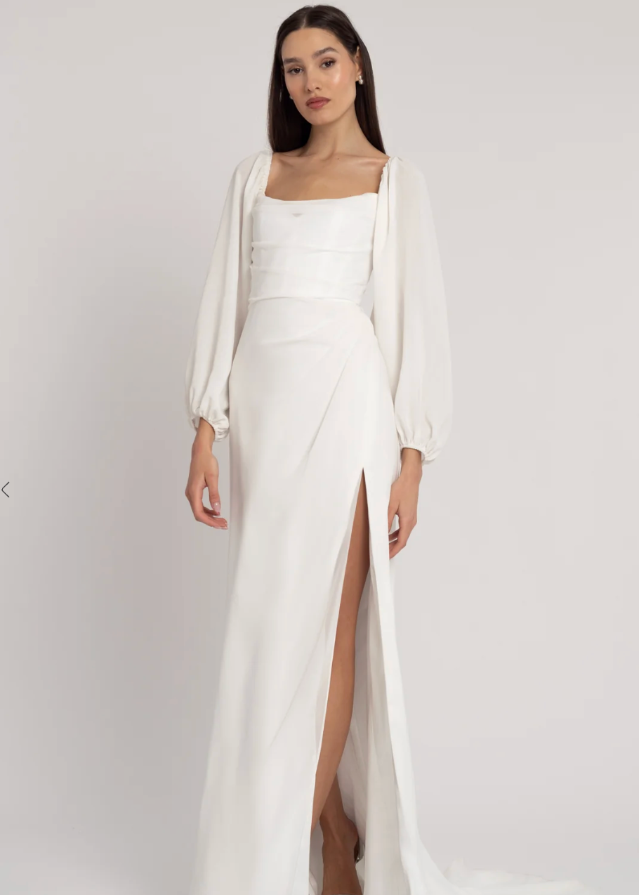 A woman modeling a floor-length white gown with a high thigh slit, square neckline, long sheer balloon sleeves, and a small train against a light gray studio background.