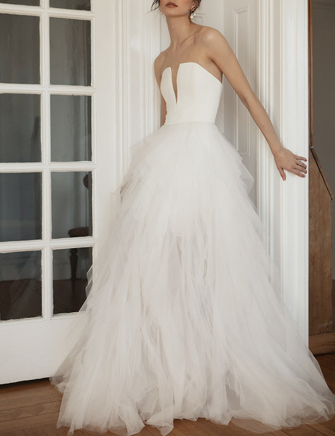 A woman wearing a strapless white wedding gown with a structured bodice and a voluminous layered tulle skirt, standing indoors near a white paneled door. Ines by Ouma