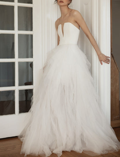 A woman wearing a strapless white wedding gown with a structured bodice and a voluminous layered tulle skirt, standing indoors near a white paneled door. Ines by Ouma