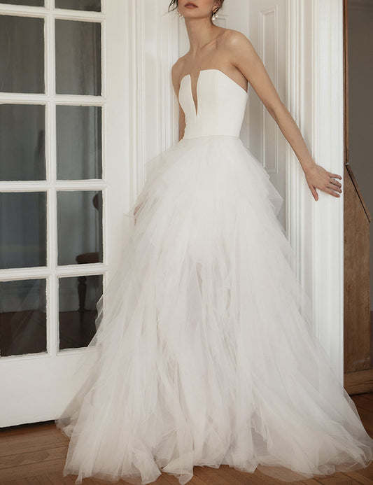 A woman wearing a strapless white wedding gown with a structured bodice and a voluminous layered tulle skirt, standing indoors near a white paneled door. Ines by Ouma