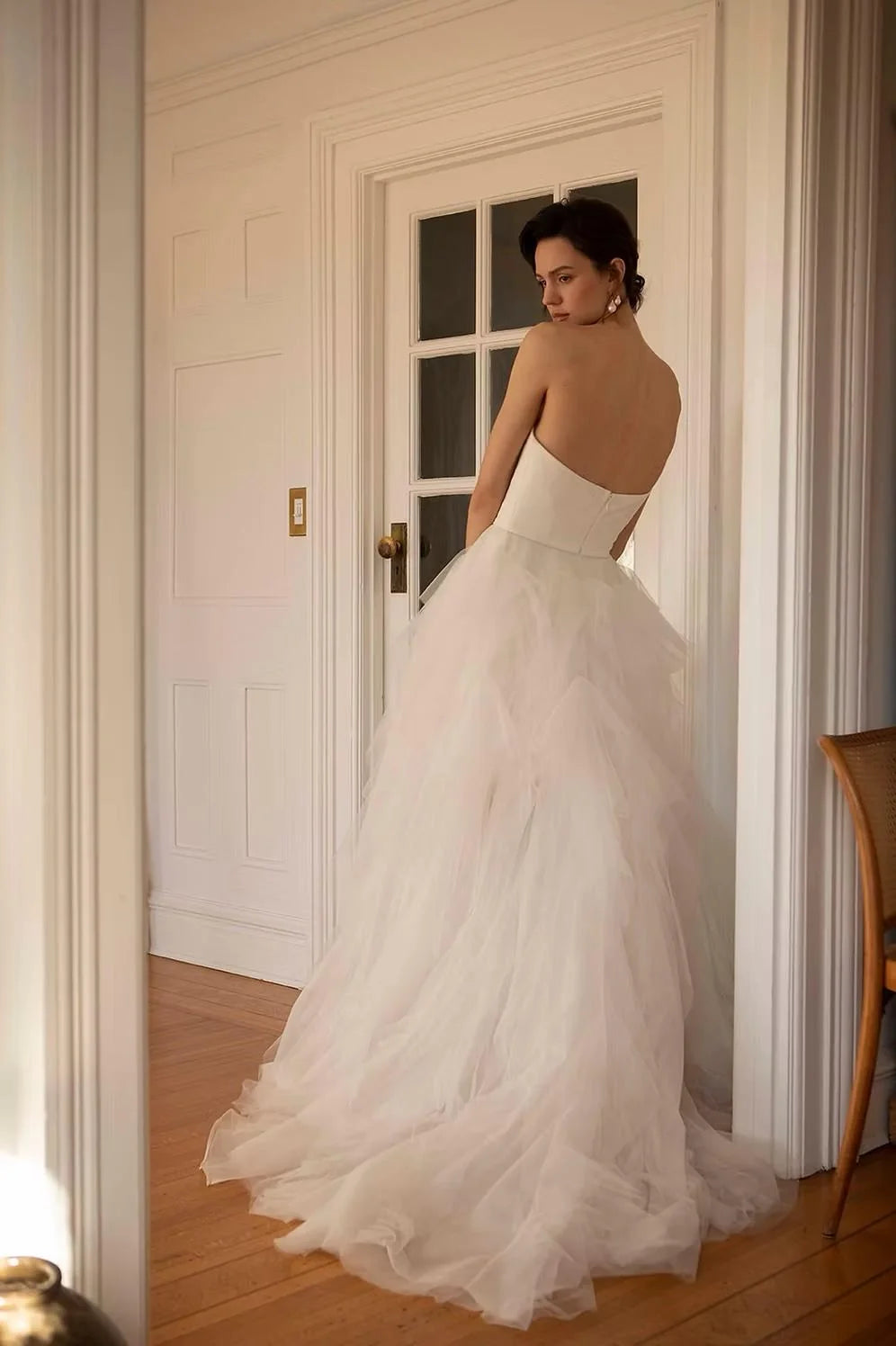 A woman wearing a strapless white tulle wedding gown stands in a doorway, viewed from behind to show the back of the dress and full train in a bright indoor room with wood floors.  Ines by Ouma