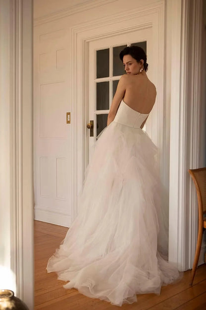 A woman wearing a strapless white tulle wedding gown stands in a doorway, viewed from behind to show the back of the dress and full train in a bright indoor room with wood floors.  Ines by Ouma