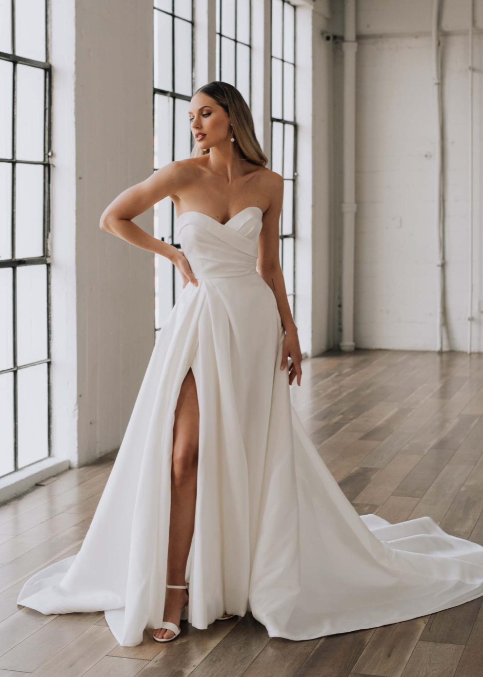 Woman in a strapless white wedding dress with a high slit standing in a room with large windows.