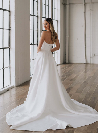 Woman in a white wedding dress standing in a room with large windows and wooden floors.