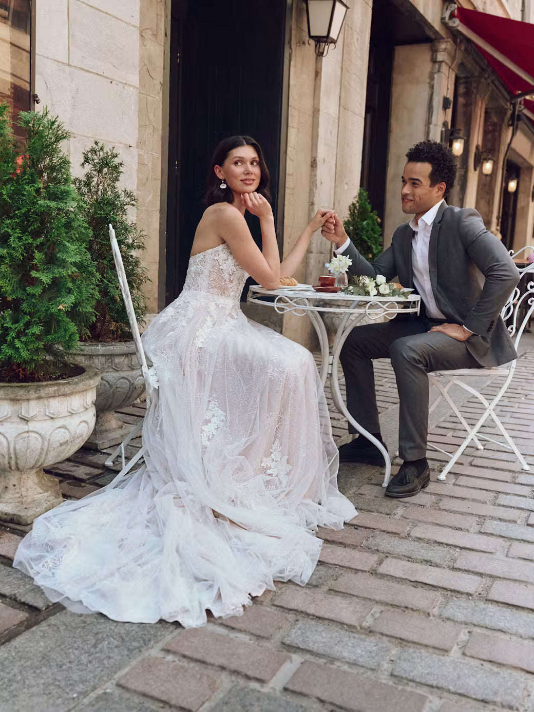A bride in a strapless white lace wedding gown sits at an outdoor cafe table holding hands with a man in a gray suit on a cobblestone street.