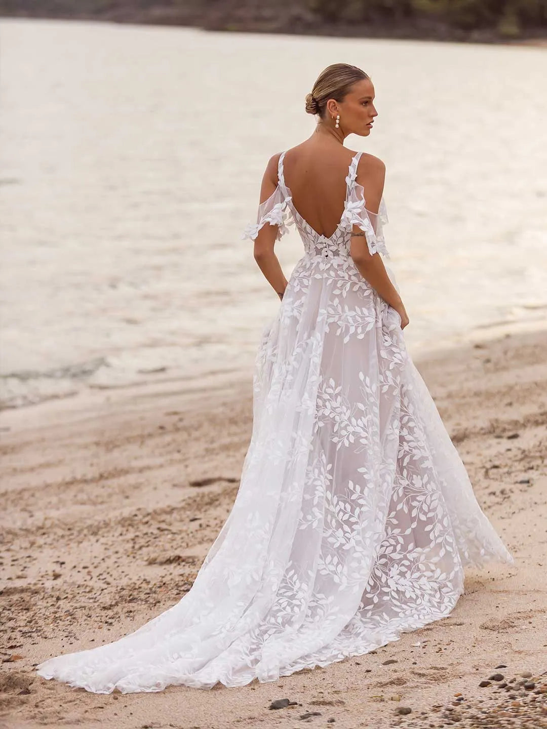 Woman in a white lace wedding dress standing on a sandy beach.