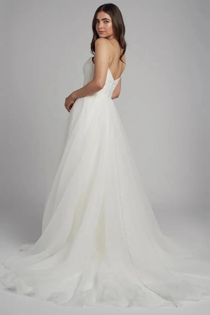 The Bronte bridal gown is cut in our lighter than air pebbled organza fabric. The flattering scoop neck is accented by the beautifully pleated bodice and elegantly draped sleeves which can be removed to create a strapless look. The full A line skirt lends a whimsical yet dramatic feel to this timeless gown.