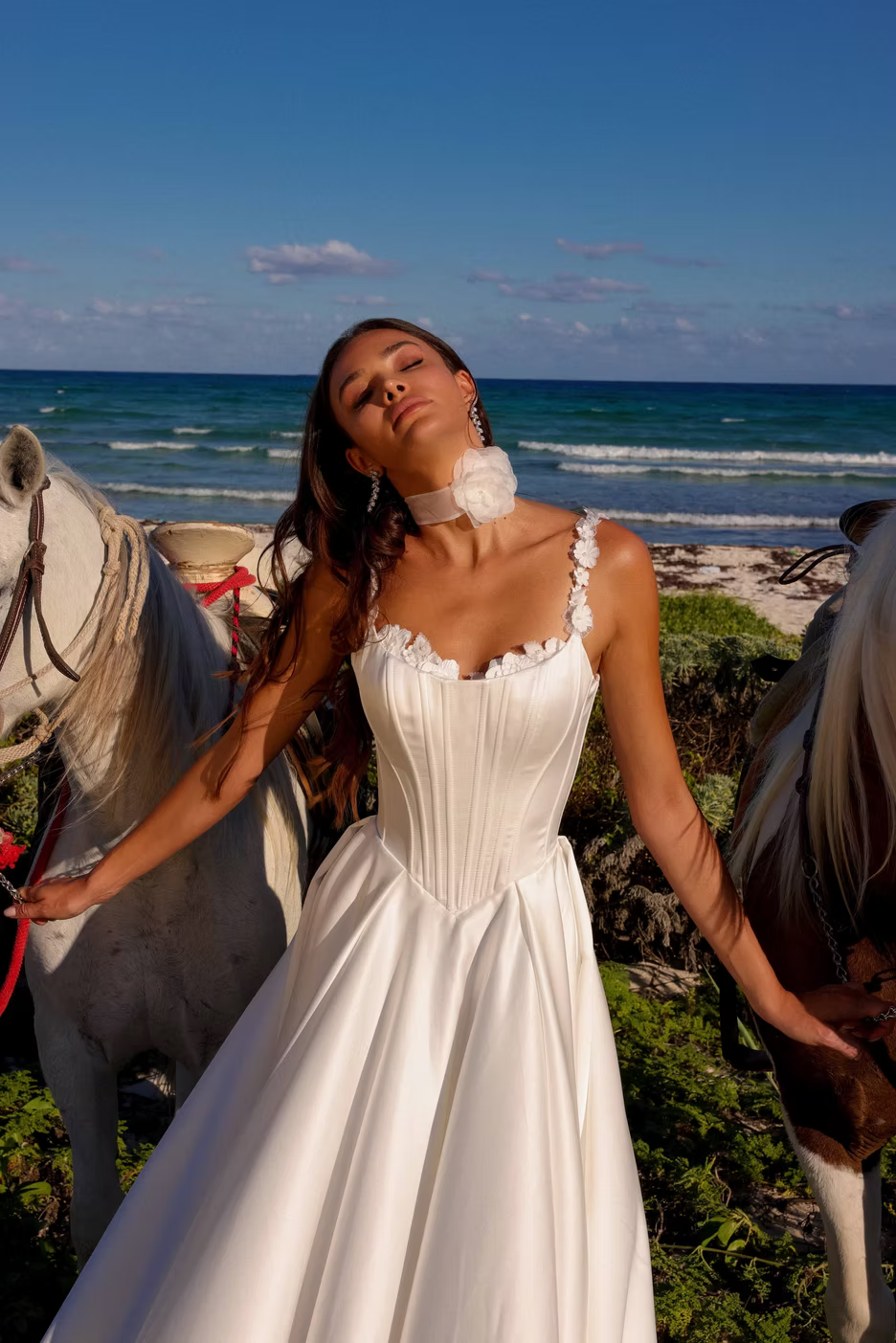 A woman wearing a white sleeveless wedding gown stands outdoors between two horses near the ocean, with blue sky and waves in the background.