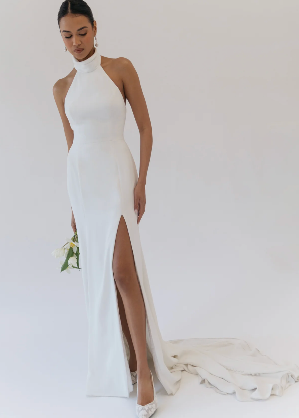A woman models a high-neck, sleeveless white wedding gown with a thigh-high front slit and a long train, standing against a plain light background and holding a small bouquet of white flowers.