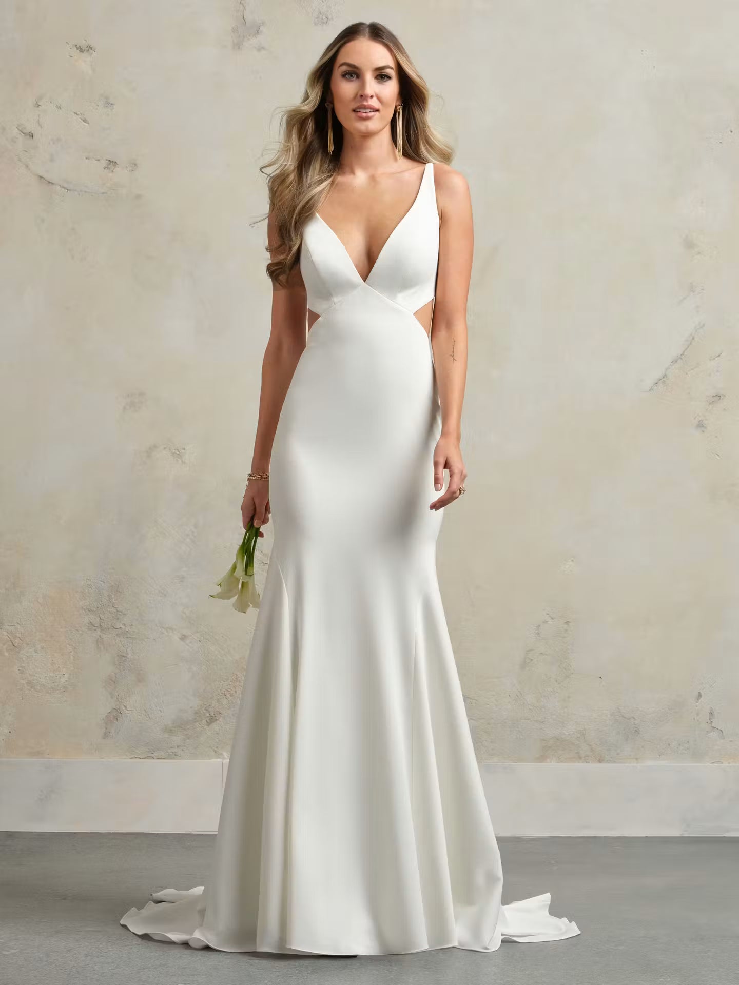 A woman modeling a floor-length white satin gown with a deep V-neckline, side cutouts, and a slight train, standing against a textured beige wall.