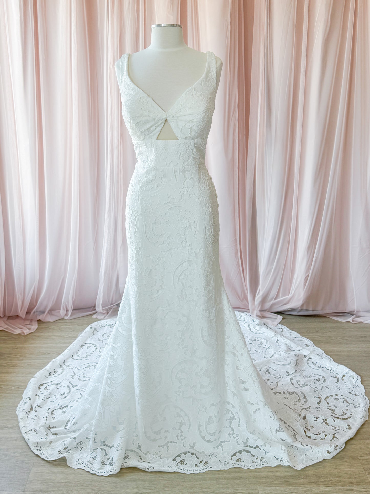 White lace mermaid-style wedding gown displayed on a mannequin with a long train, set against soft pink draped curtains and a light wood floor at Never Knew I Needed
