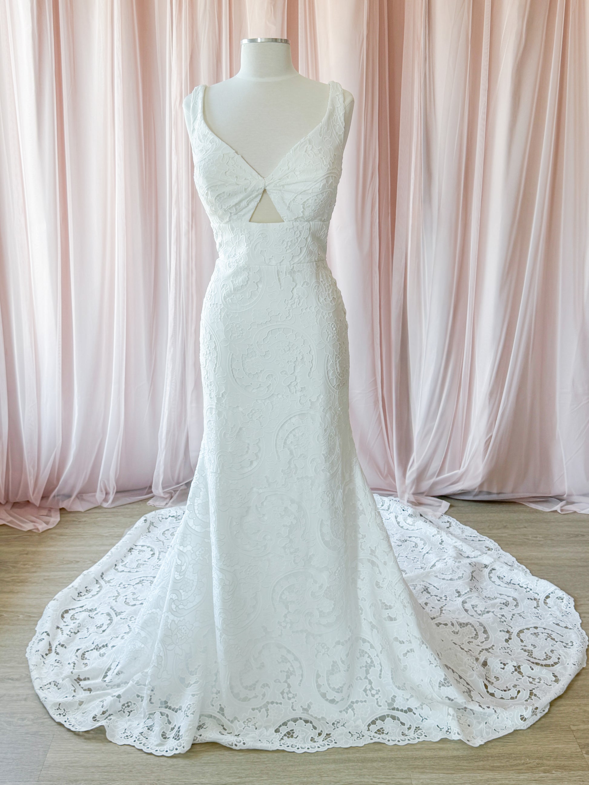 White lace mermaid-style wedding gown displayed on a mannequin with a long train, set against soft pink draped curtains and a light wood floor at Never Knew I Needed