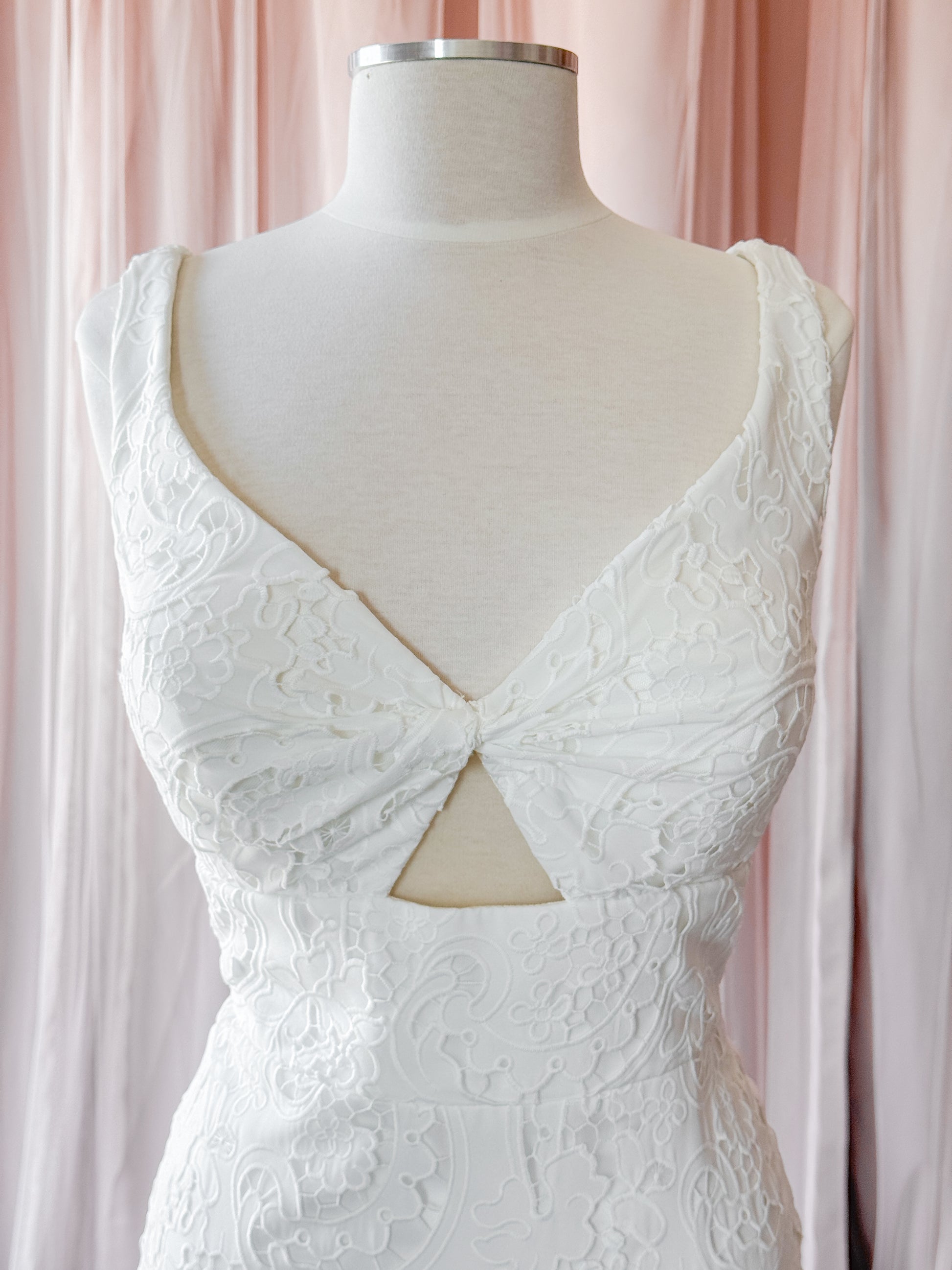 White lace sleeveless dress displayed on a cream mannequin torso, featuring a deep V-neckline with a twisted front detail and a triangular cutout at the waist, set against soft pink draped fabric in a fitting room bridal shop NKIN