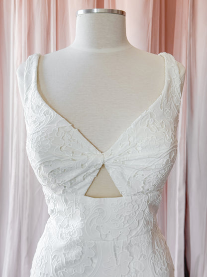 White lace sleeveless dress displayed on a cream mannequin torso, featuring a deep V-neckline with a twisted front detail and a triangular cutout at the waist, set against soft pink draped fabric in a fitting room bridal shop NKIN