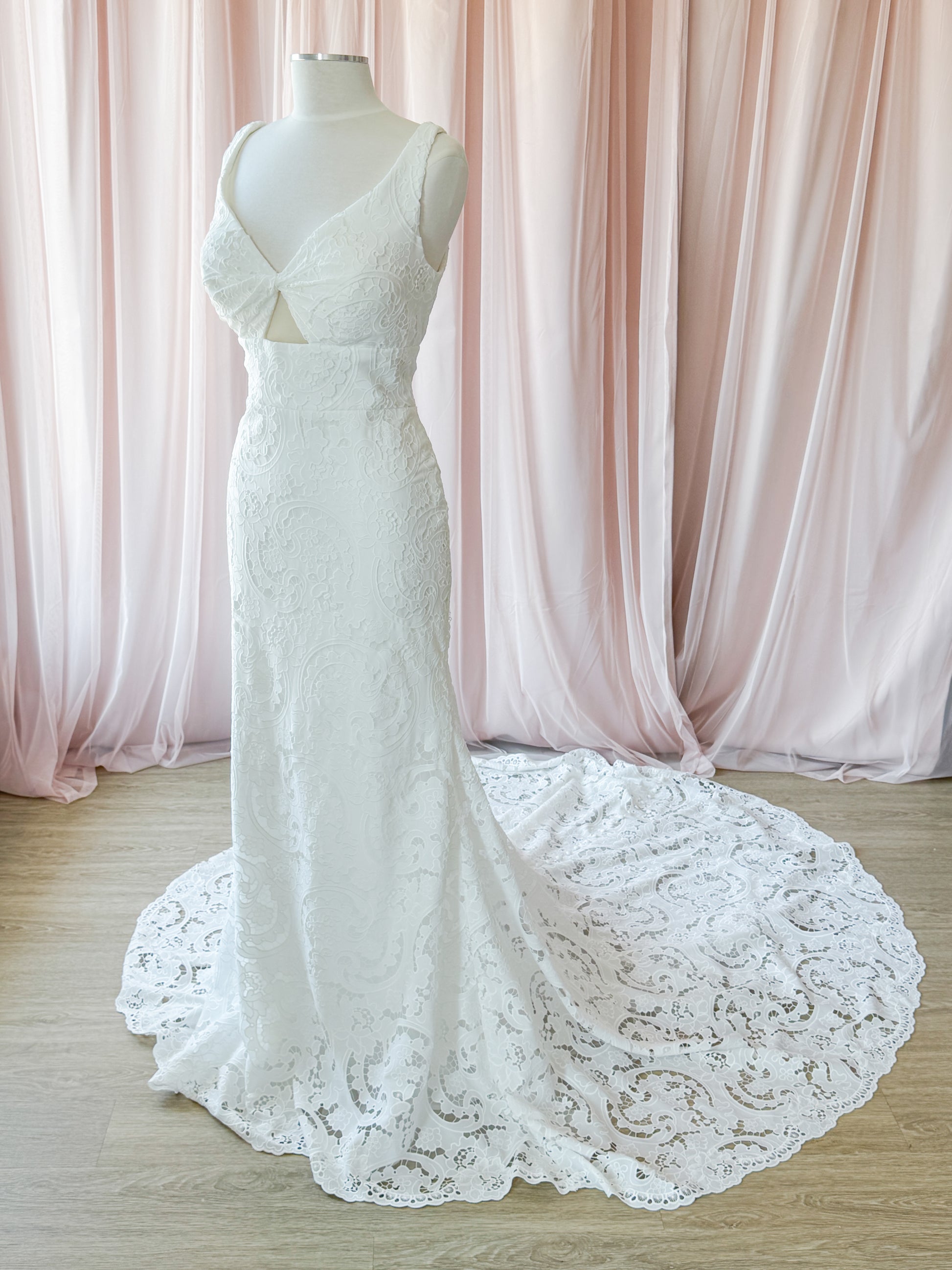 White lace column-style wedding gown displayed on a mannequin in front of soft pink draped curtains and a light wood floor. Background is a fitting room at NKIN Ottawa Bridal Outlet