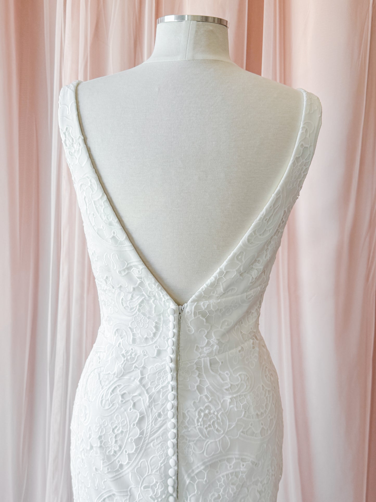 White lace wedding dress shown from the back on a cream mannequin, featuring a deep V-neckline with a button closure and floral lace detailing, set against a soft pink draped fabric background at NKIN ottawa bridal shop
