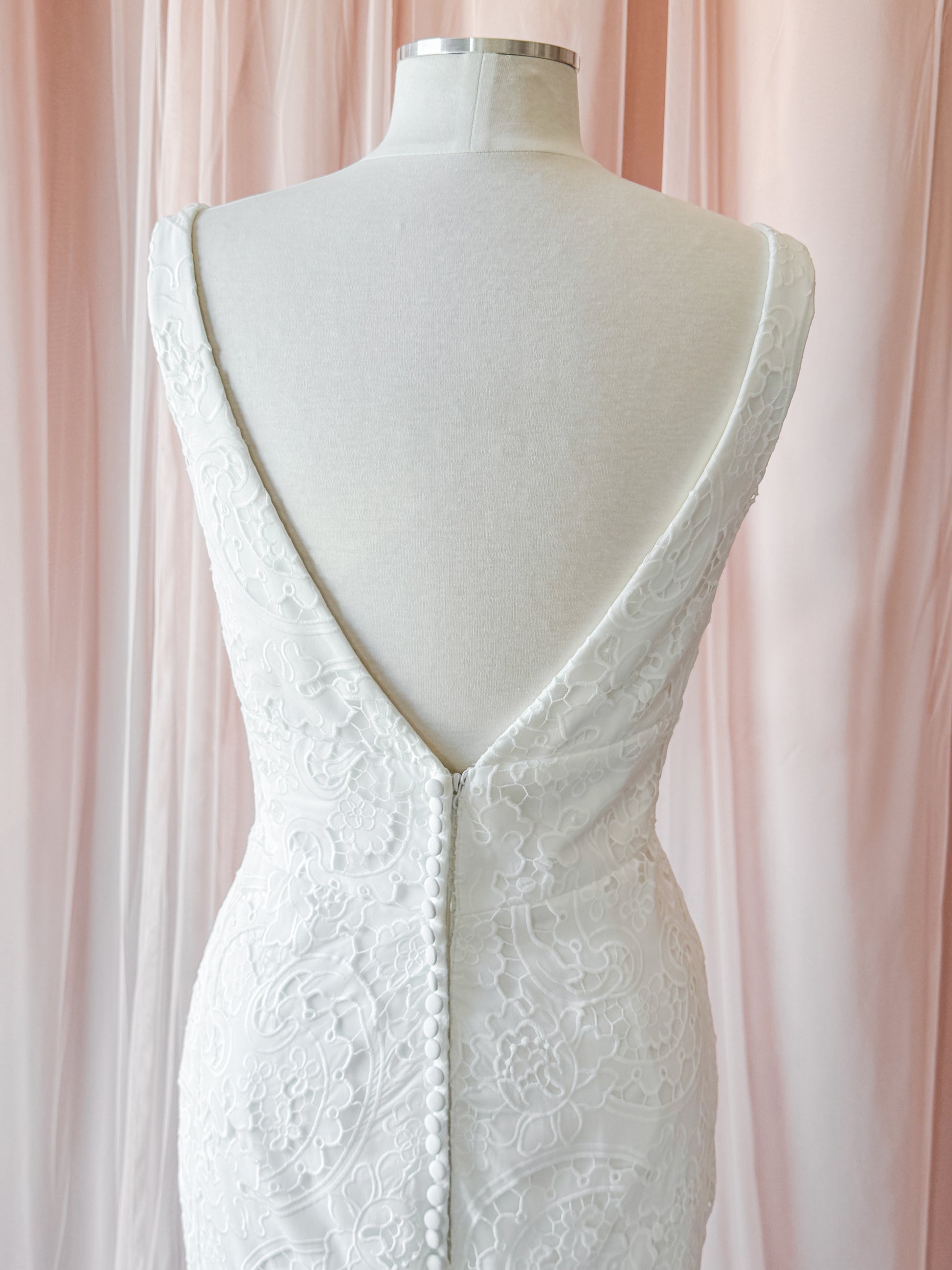White lace wedding dress shown from the back on a cream mannequin, featuring a deep V-neckline with a button closure and floral lace detailing, set against a soft pink draped fabric background at NKIN ottawa bridal shop
