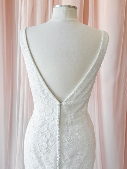 White lace wedding dress shown from the back on a cream mannequin, featuring a deep V-neckline with a button closure and floral lace detailing, set against a soft pink draped fabric background at NKIN ottawa bridal shop
