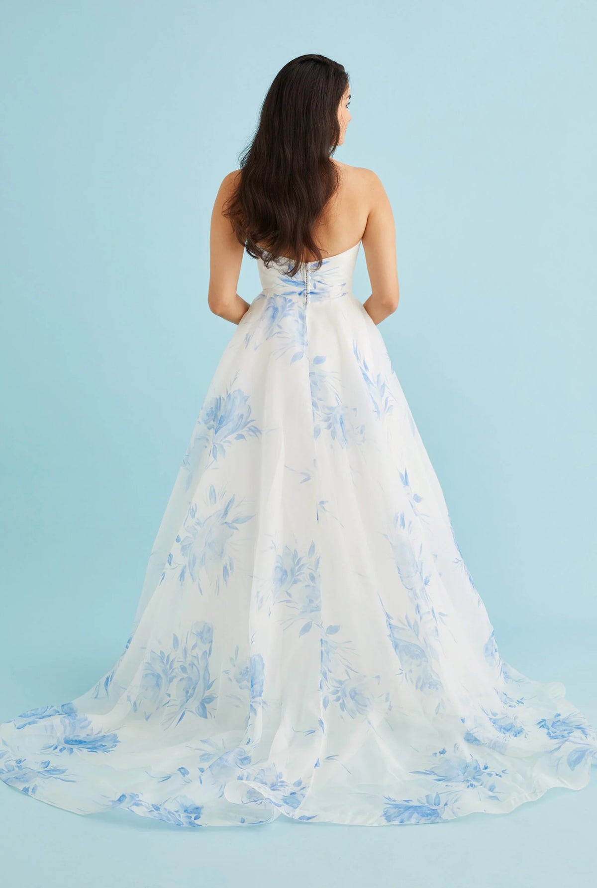 A woman viewed from behind wearing a strapless white ball gown with a blue floral pattern and a long train, set against a solid light blue studio background.