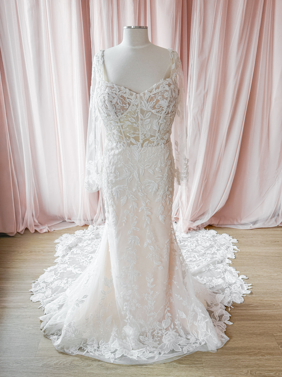 A white lace wedding gown displayed on a mannequin, featuring a sweetheart neckline, long sheer sleeves, and a dramatic flared lace train, set against a backdrop of soft pink draped fabric and a light wood floor.