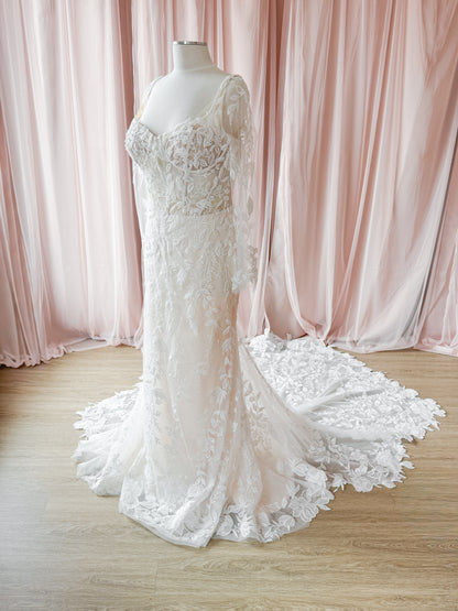 An ivory lace wedding gown with long sheer sleeves and a dramatic train, displayed on a mannequin in front of soft pink draped curtains and a light wood floor.