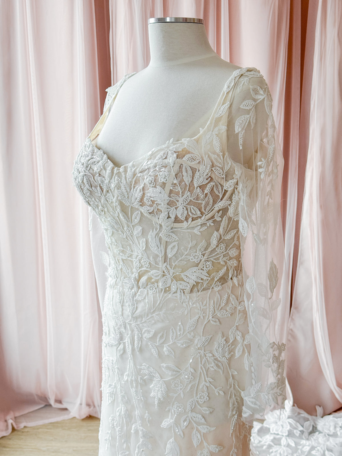 An ivory lace wedding dress displayed on a cream mannequin torso, set against a soft pink draped fabric backdrop.
