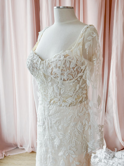 An ivory lace wedding dress displayed on a cream mannequin torso, set against a soft pink draped fabric backdrop.