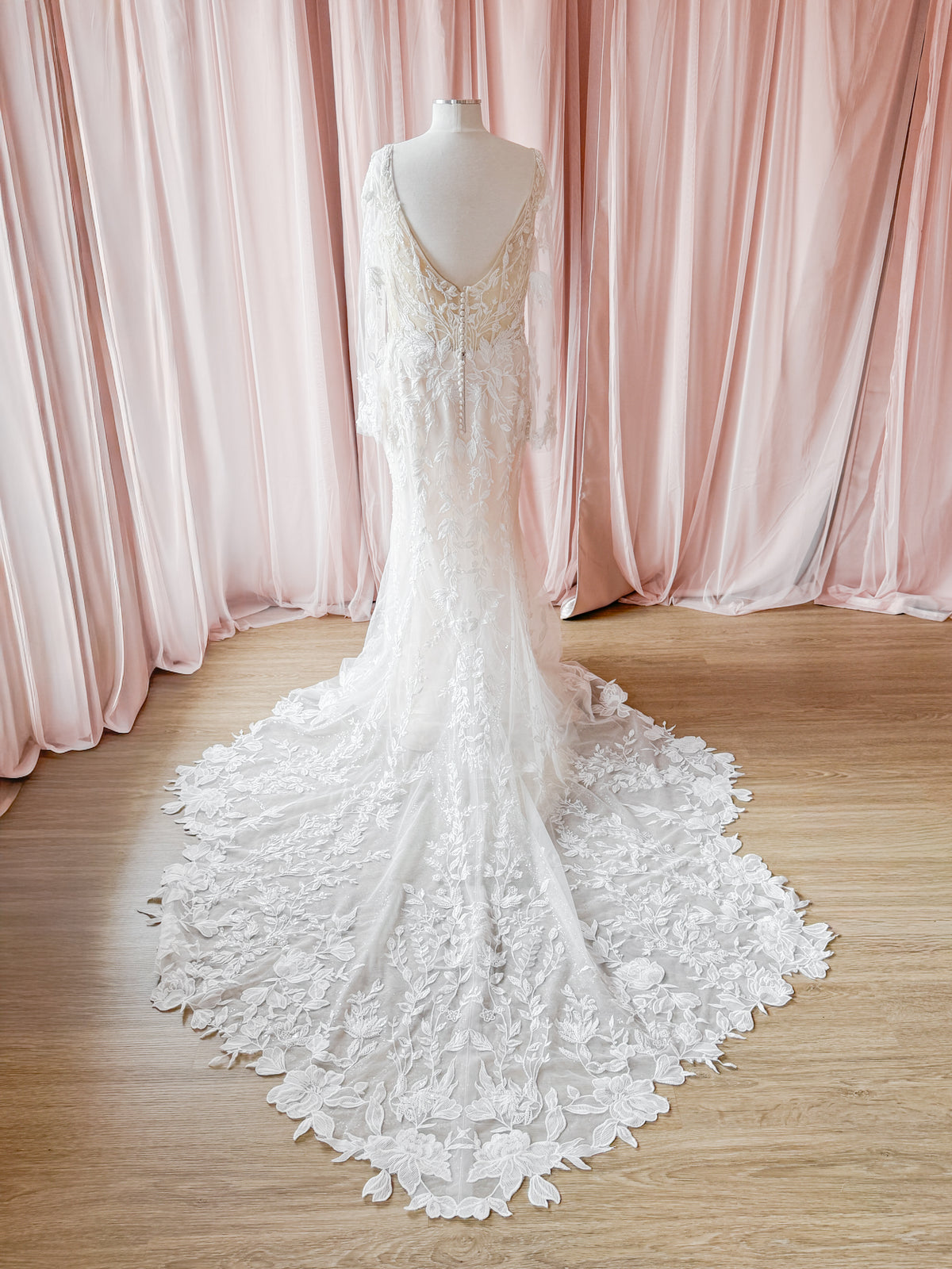 An ivory lace wedding gown with a long train is displayed on a mannequin in front of soft pink draped curtains and a light wood floor.