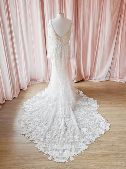 An ivory lace wedding gown with a long train is displayed on a mannequin in front of soft pink draped curtains and a light wood floor.