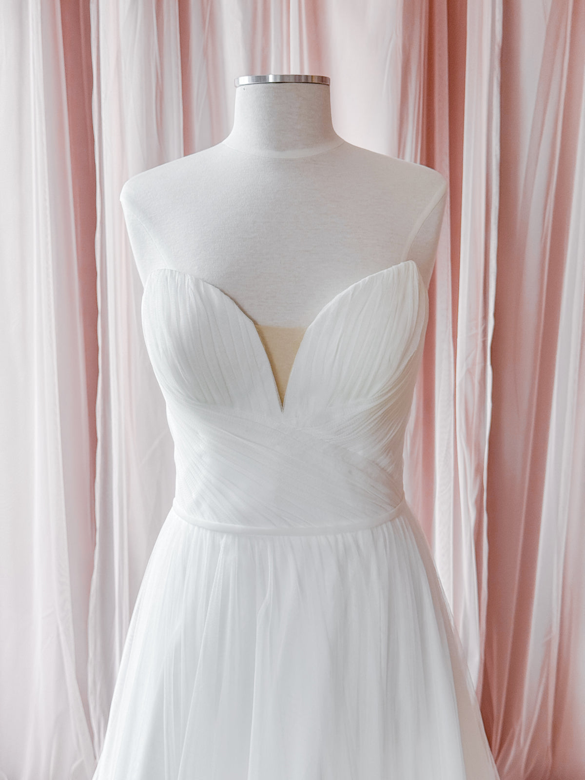 A white strapless sweetheart dress displayed on a white mannequin torso, set against a backdrop of soft pink and white draped fabric curtains.