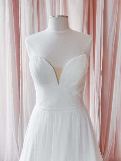 A white strapless sweetheart dress displayed on a white mannequin torso, set against a backdrop of soft pink and white draped fabric curtains.