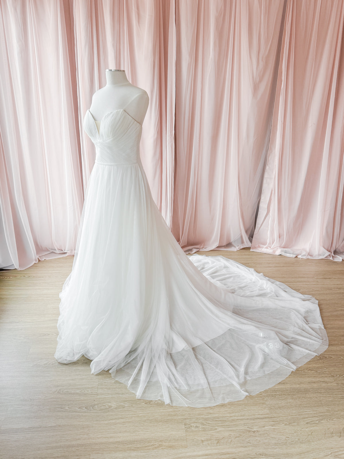 A white strapless wedding gown displayed on a mannequin, featuring a sweetheart neckline, fitted bodice, and a voluminous layered tulle skirt with a long train, set against a backdrop of draped blush pink curtains and light wood flooring.