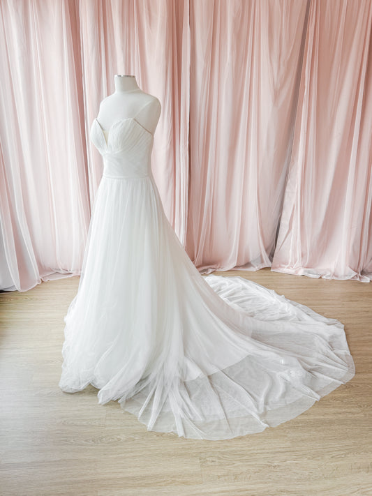A white strapless wedding gown displayed on a mannequin, featuring a sweetheart neckline, fitted bodice, and a voluminous layered tulle skirt with a long train, set against a backdrop of draped blush pink curtains and light wood flooring.