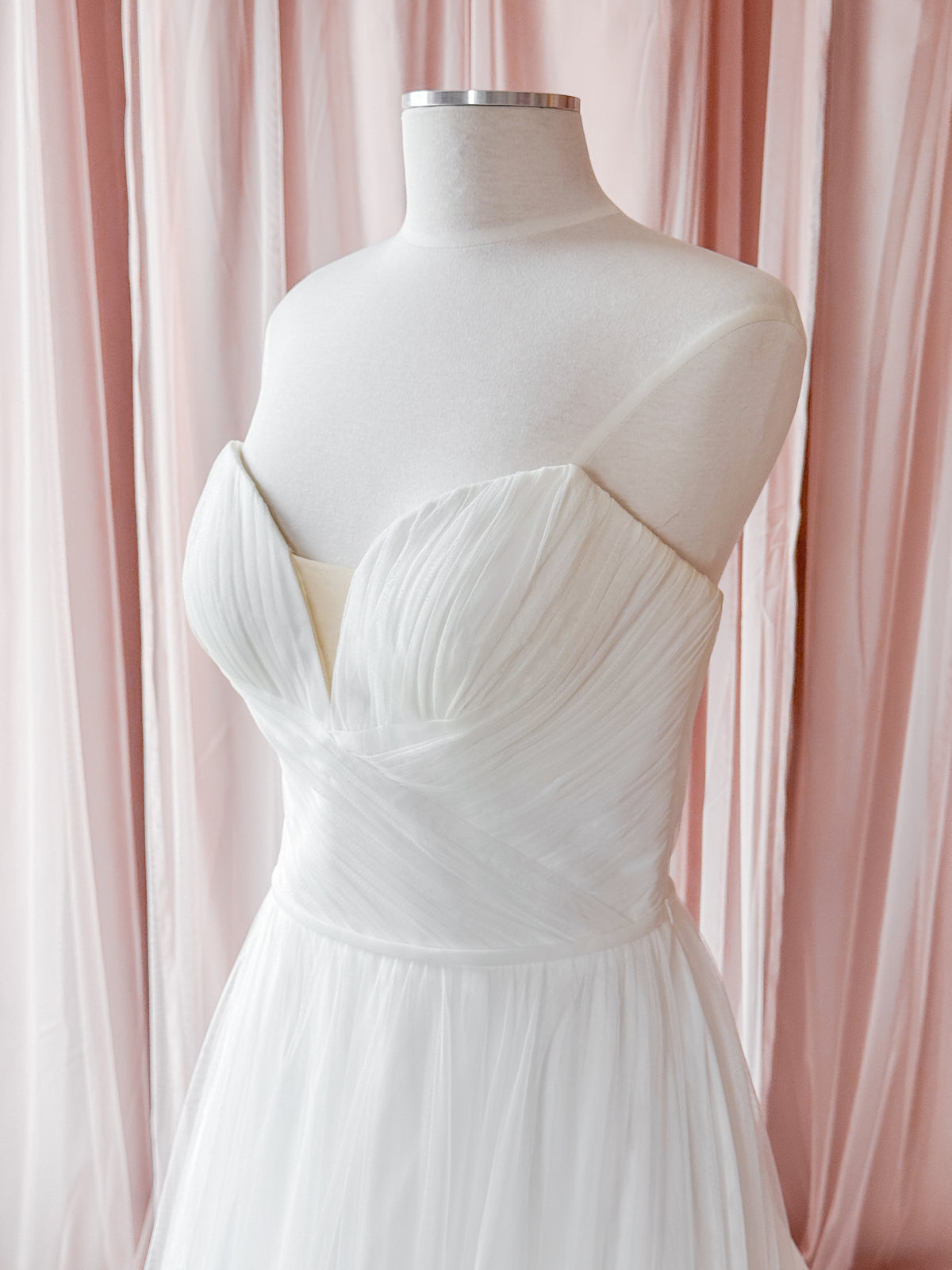 A white strapless wedding dress with a sweetheart neckline and pleated bodice, displayed on a white mannequin dress form against a soft pink draped fabric backdrop.