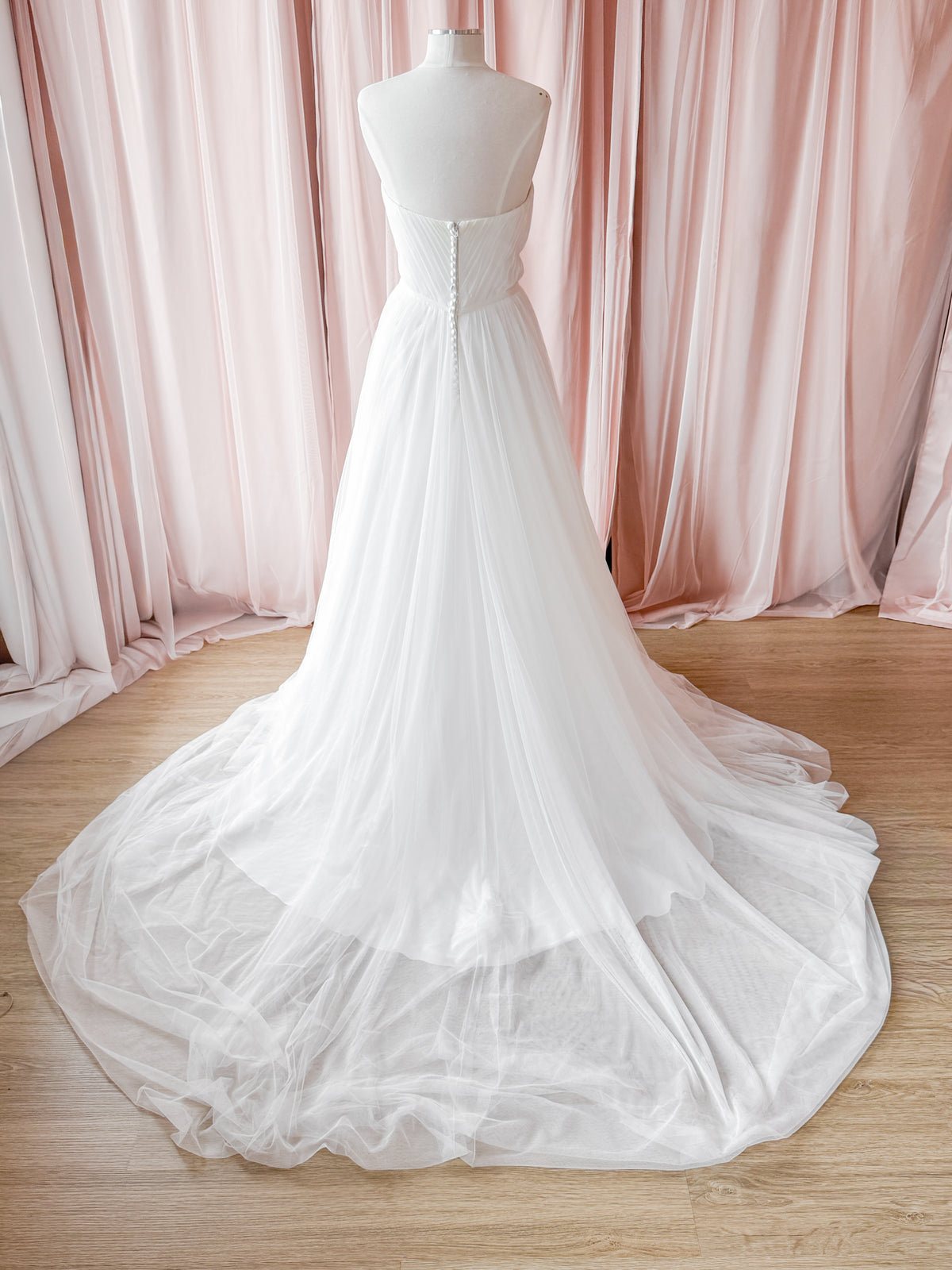 A white strapless wedding gown displayed on a mannequin, featuring a structured lace-up back bodice and a voluminous tulle skirt with a long train, set against a backdrop of soft pink draped fabric and a light wood floor.