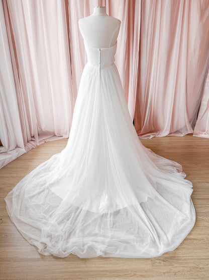 A white strapless wedding gown displayed on a mannequin, featuring a structured lace-up back bodice and a voluminous tulle skirt with a long train, set against a backdrop of soft pink draped fabric and a light wood floor.