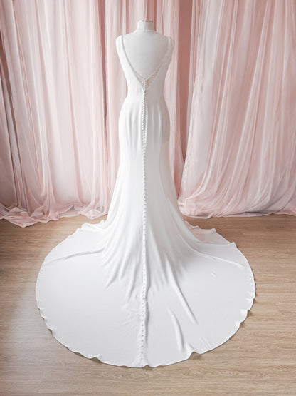 A white sleeveless wedding dress displayed on a mannequin from the back, featuring a deep V-neckline, a beaded button back, and a long flowing train, set against a backdrop of soft pink draped fabric and a light wood floor.