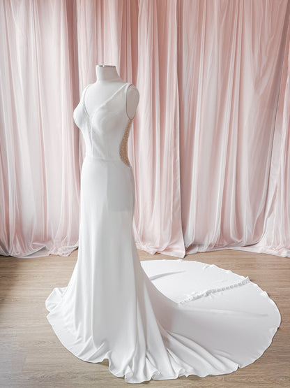 A sleeveless white crepe minimal beaded wedding gown displayed on a mannequin against a backdrop of soft pink draped curtains and a light wood floor.