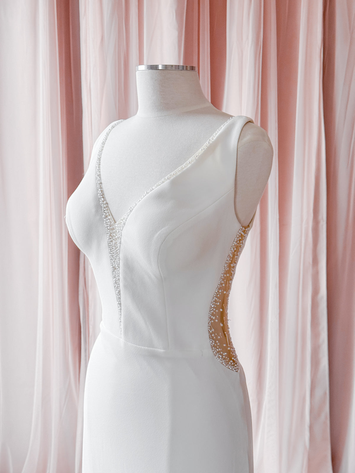 A white sleeveless gown displayed on a white mannequin torso, featuring a deep V-neckline with beaded trim and a curved beaded side accent, set against a soft pink draped fabric backdrop.