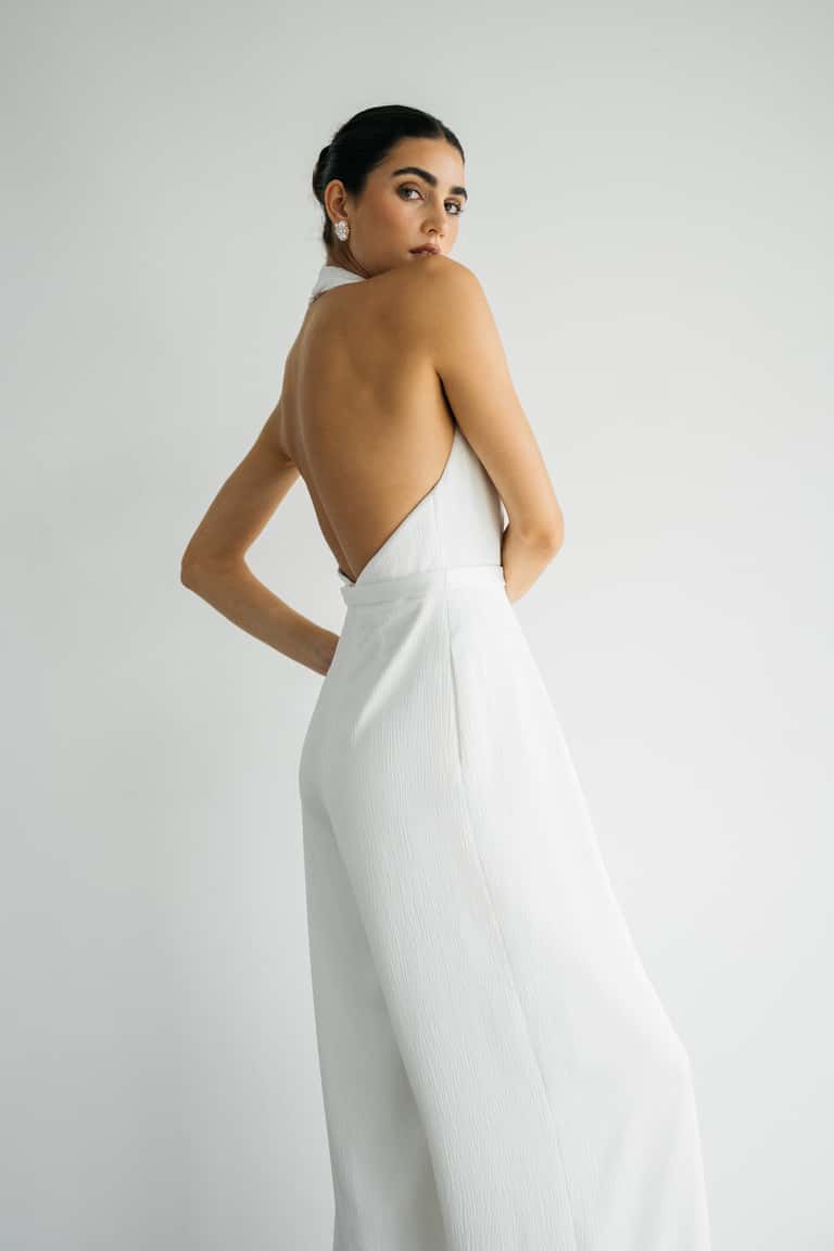 Woman wearing a white wedding jumpsuit with an open back against a plain background