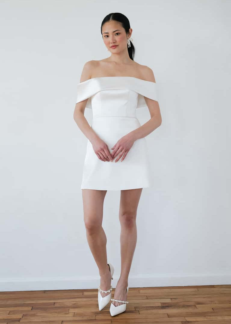 Woman wearing a white off-shoulder dress against a plain white wall.