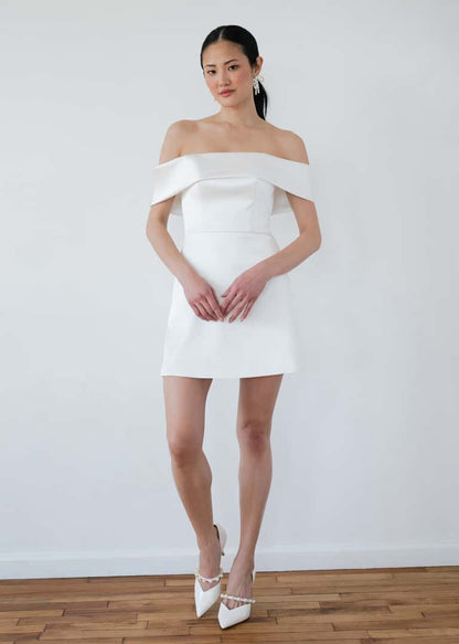 Woman wearing a white off-shoulder dress against a plain white wall.