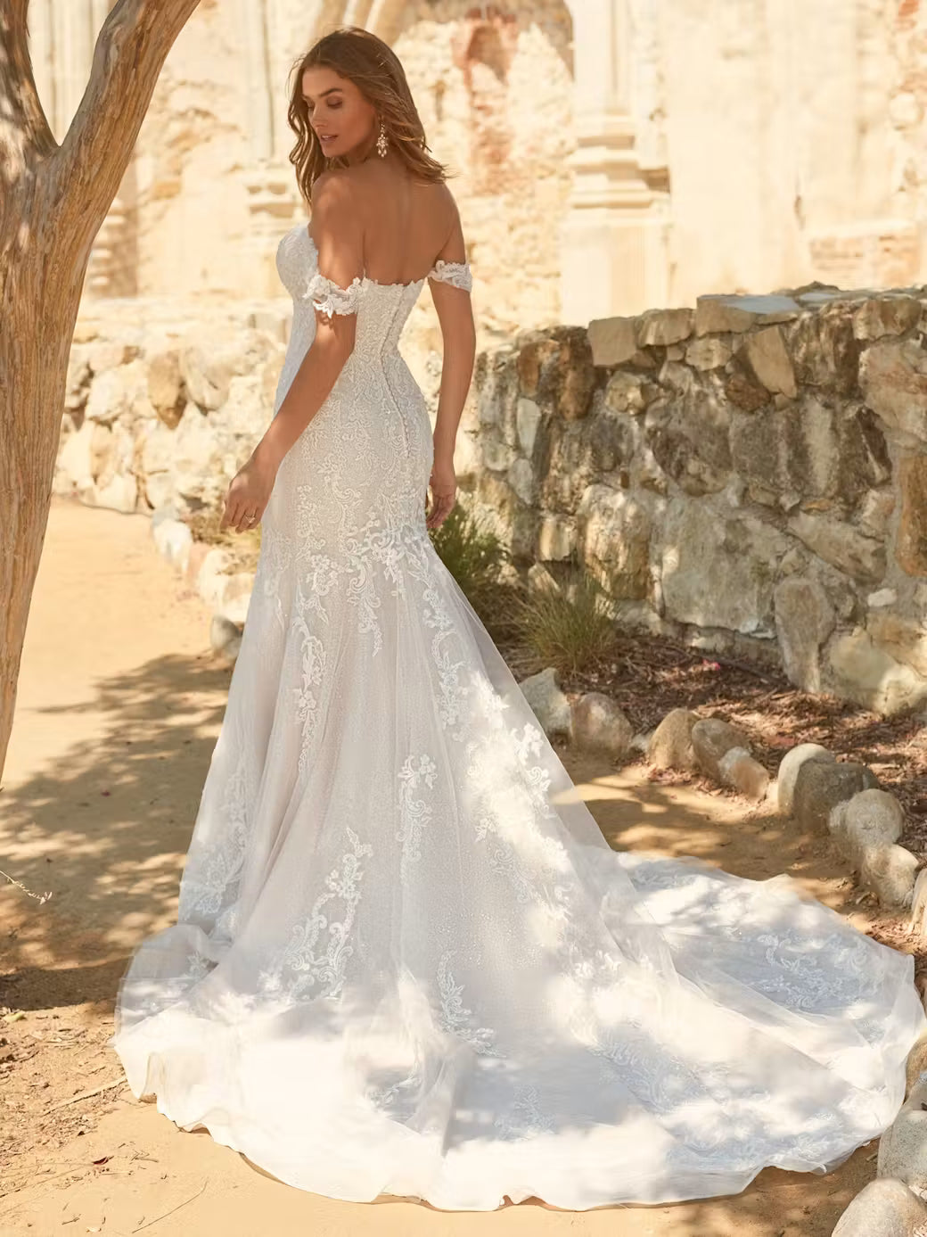 Woman in a white lace wedding dress, frederique by maggie sottero, standing outdoors with stone walls in the background