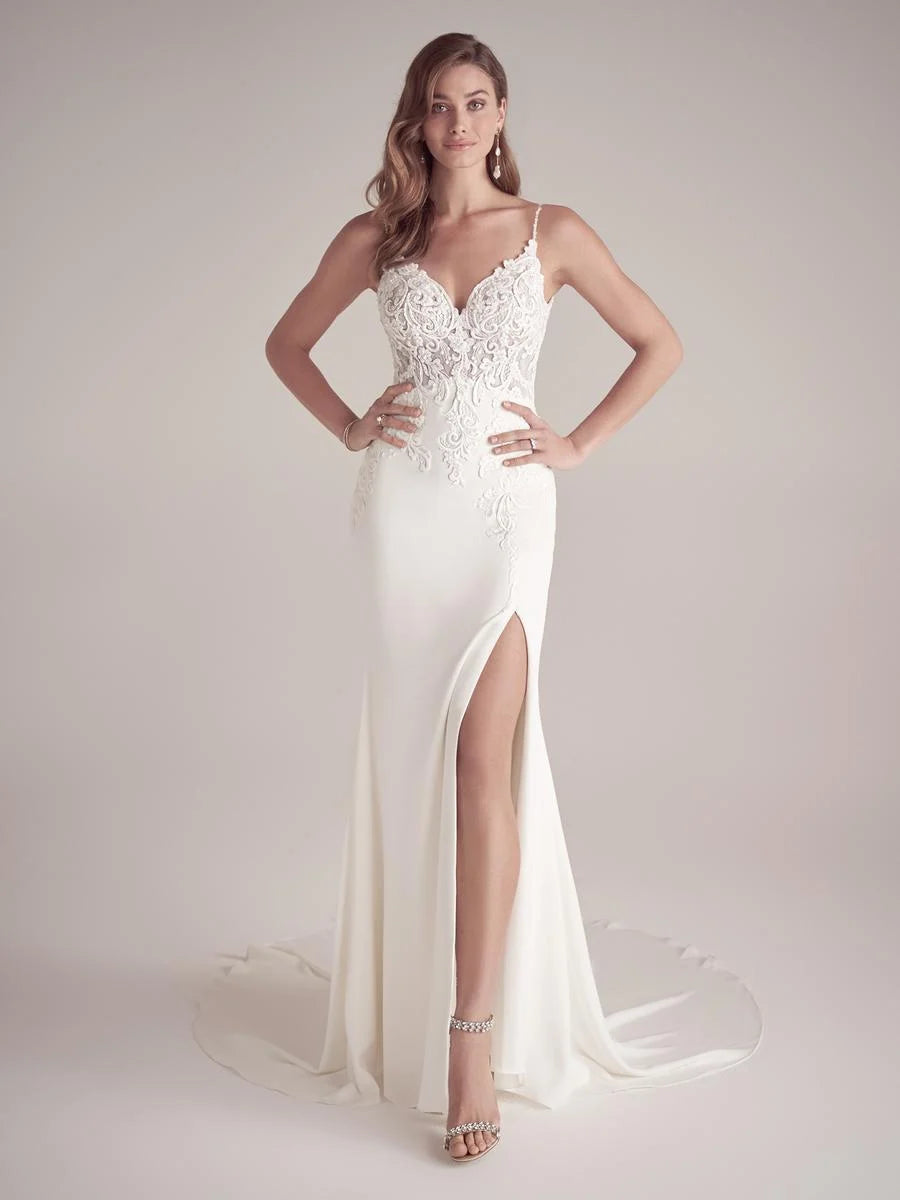A woman modeling a floor-length white wedding gown with thin straps, a lace-embroidered bodice, a high thigh slit, and a short train against a plain light beige studio background. Available at NKIN