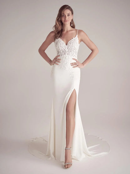 A woman modeling a floor-length white wedding gown with thin straps, a lace-embroidered bodice, a high thigh slit, and a short train against a plain light beige studio background. Available at NKIN